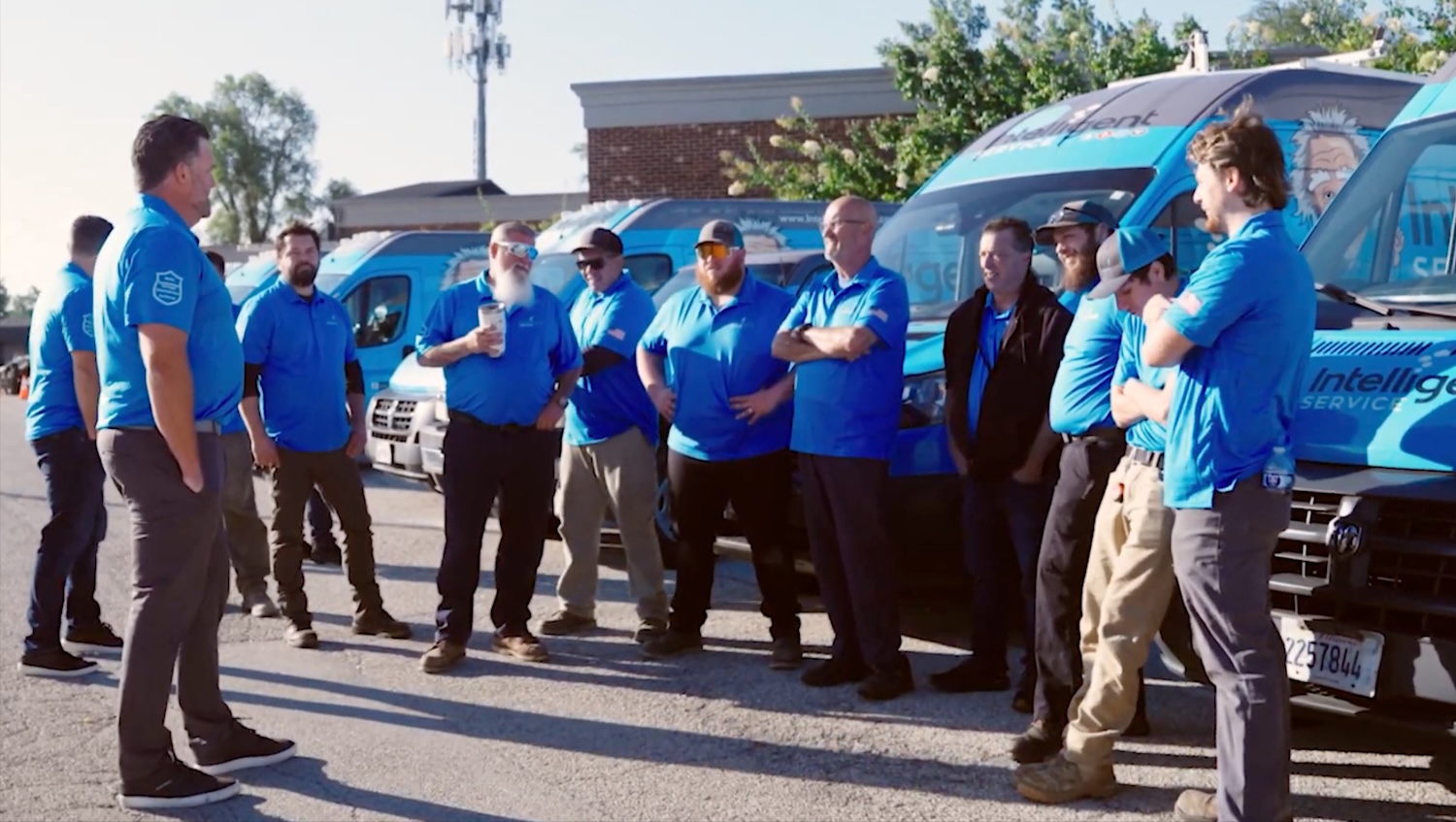 Team of technicians in blue shirts stands by service vans, gathered outdoors for briefing.