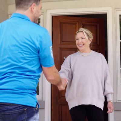 Smiling woman greets repairman at her front door, shaking hands in a friendly exchange.