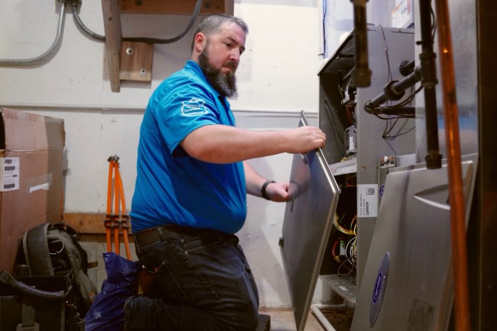 Technician servicing HVAC system in utility room, kneeling and inspecting internal components for maintenance.