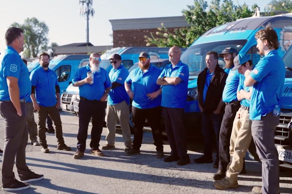 Team of technicians in blue shirts stands by service vans, gathered outdoors for briefing.