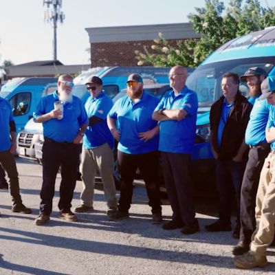 Team of technicians in blue shirts stands by service vans, gathered outdoors for briefing.